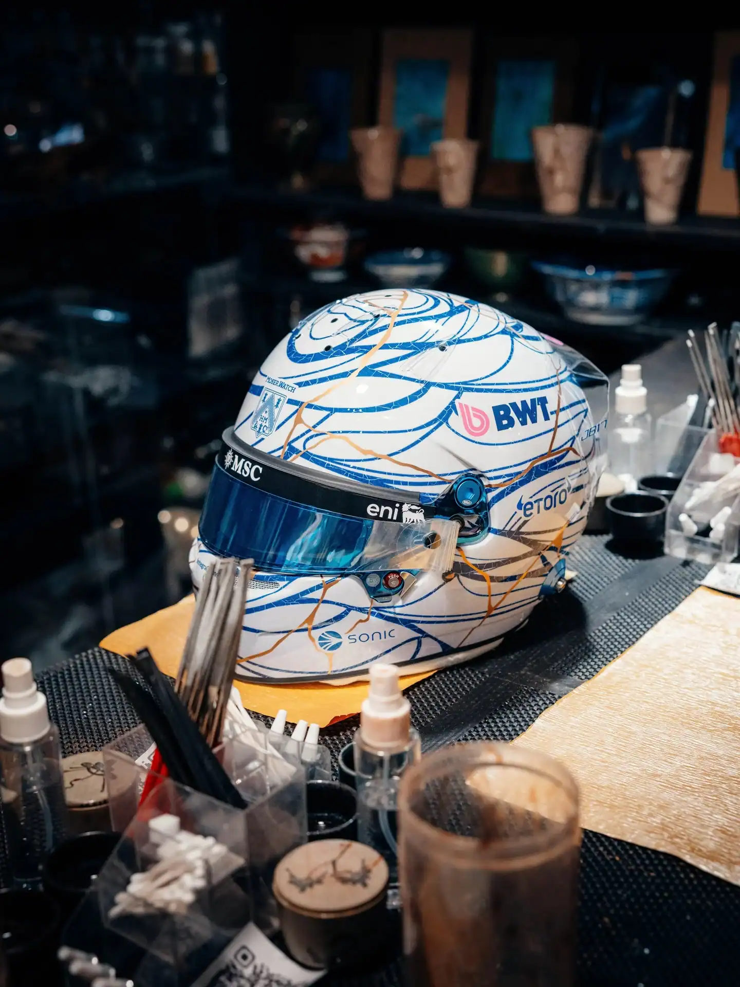 Pierre Gaslys Japan-GP-2026-Helm ausgestellt in einer Kintsugi-K&uuml;nstlerwerkstatt mit traditionellen Reparaturwerkzeugen &mdash; BWT-Alpine-Sponsoren sichtbar auf wei&szlig;-blauem Design