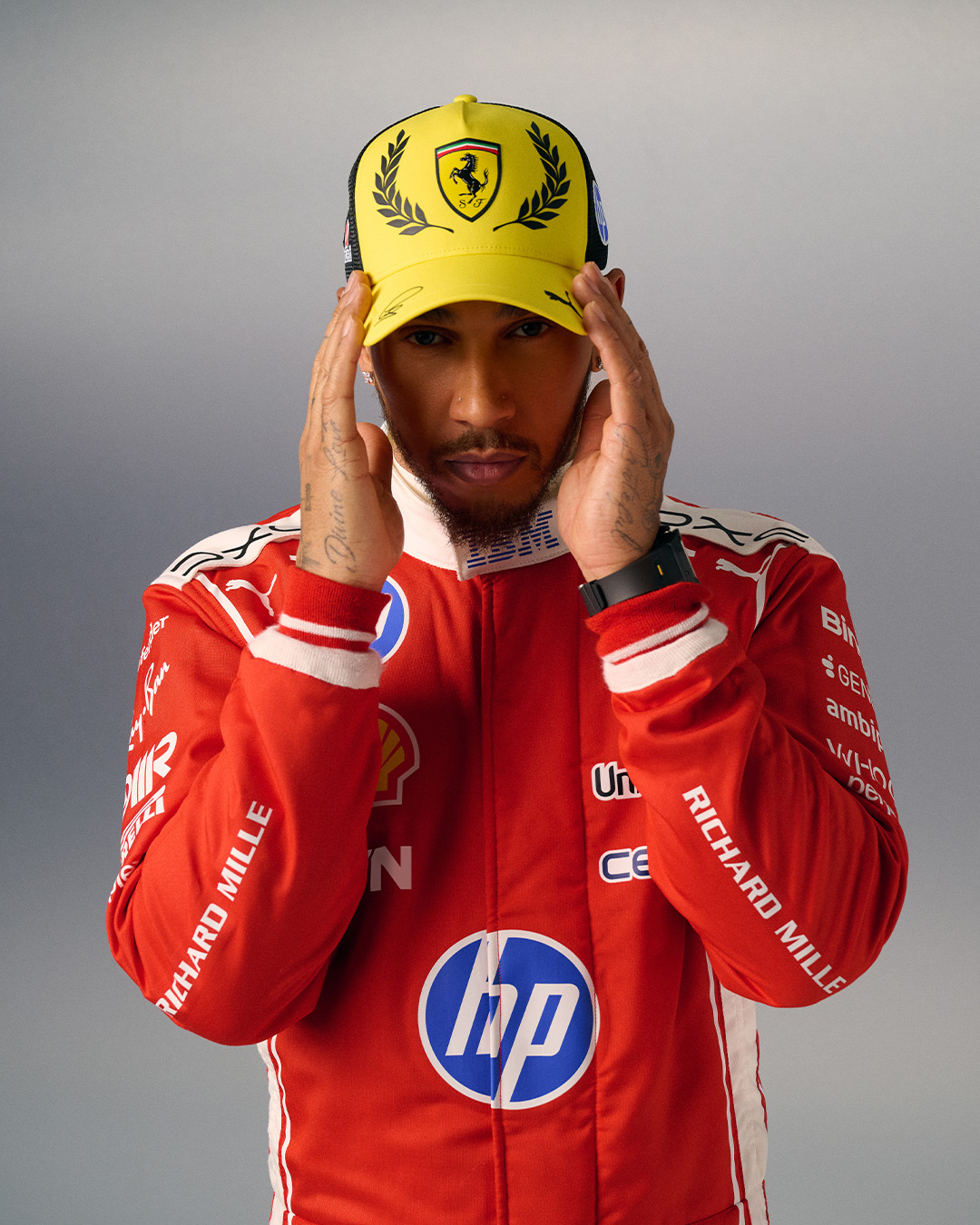 Lewis Hamilton adjusts a yellow Ferrari cap while wearing the red Ferrari race suit for the 2026 British Grand Prix.