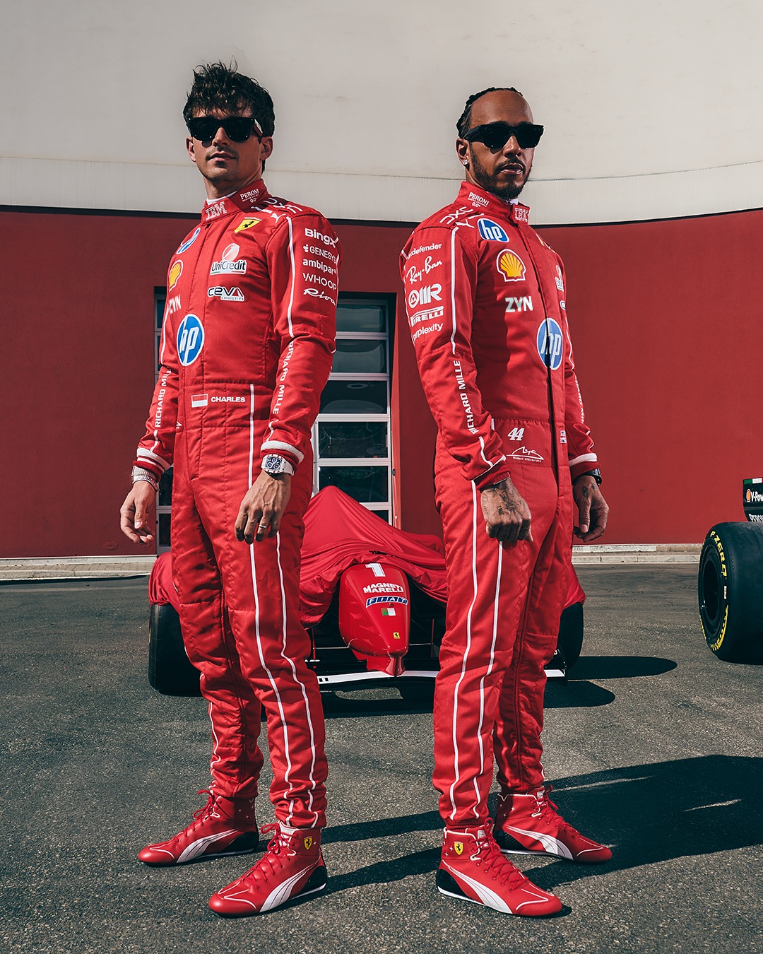 Charles Leclerc y Lewis Hamilton de pie uno al lado del otro con los trajes de carrera especiales Monza 2026 de Ferrari — rojo con una línea blanca recorriendo la longitud de los brazos y las piernas — frente a un coche cubierto cuyo ex