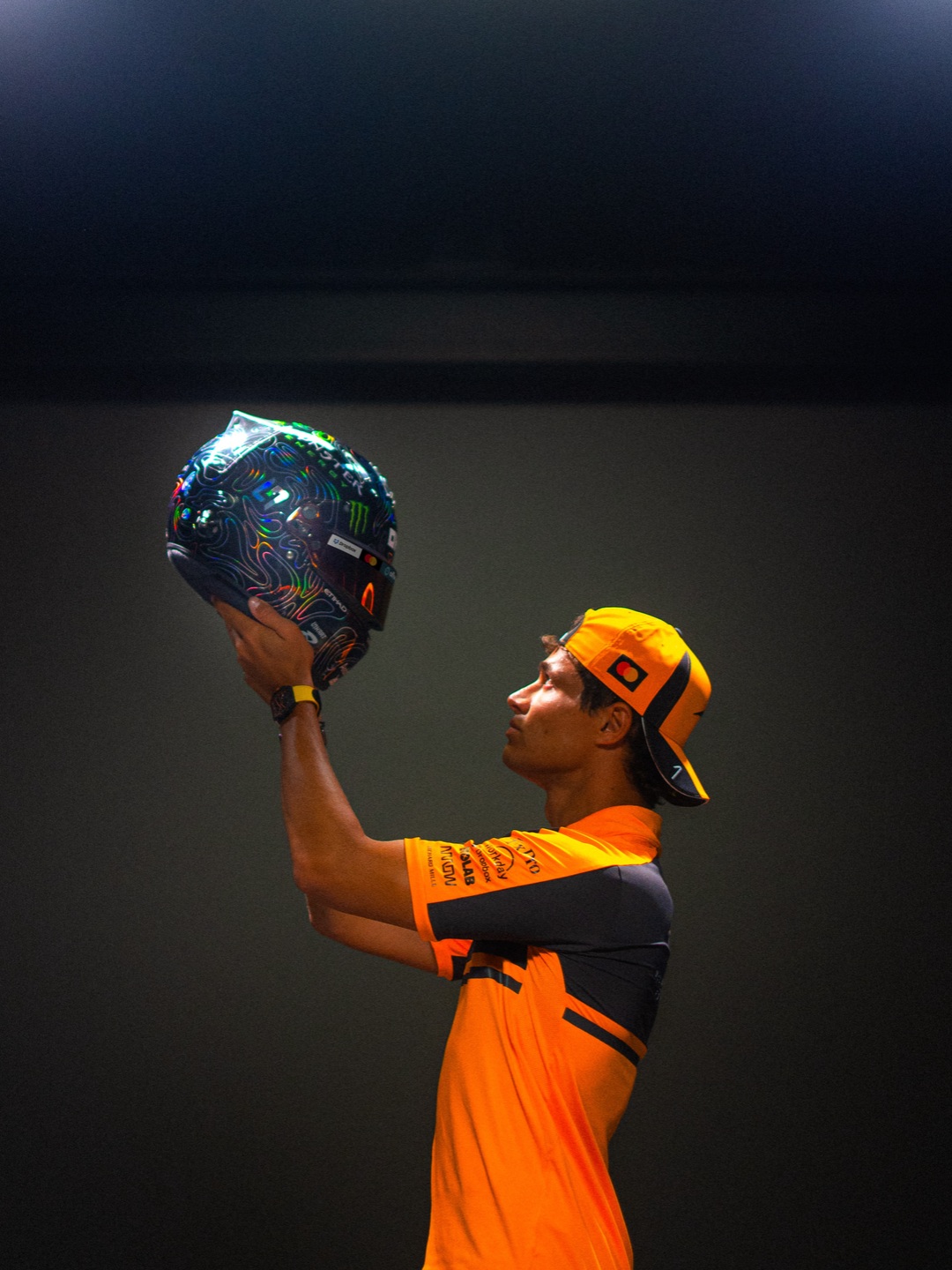 Lando Norris lifts his holographic 2026 Dutch GP helmet under studio lights in McLaren team kit