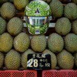Front view of Alex Albon's 2026 Singapore GP helmet, green durian-skin paint with Atlassian, Barclays and Kraken logos, on a durian stall