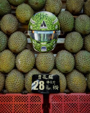 Front view of Alex Albon's 2026 Singapore GP helmet, green durian-skin paint with Atlassian, Barclays and Kraken logos, on a durian stall