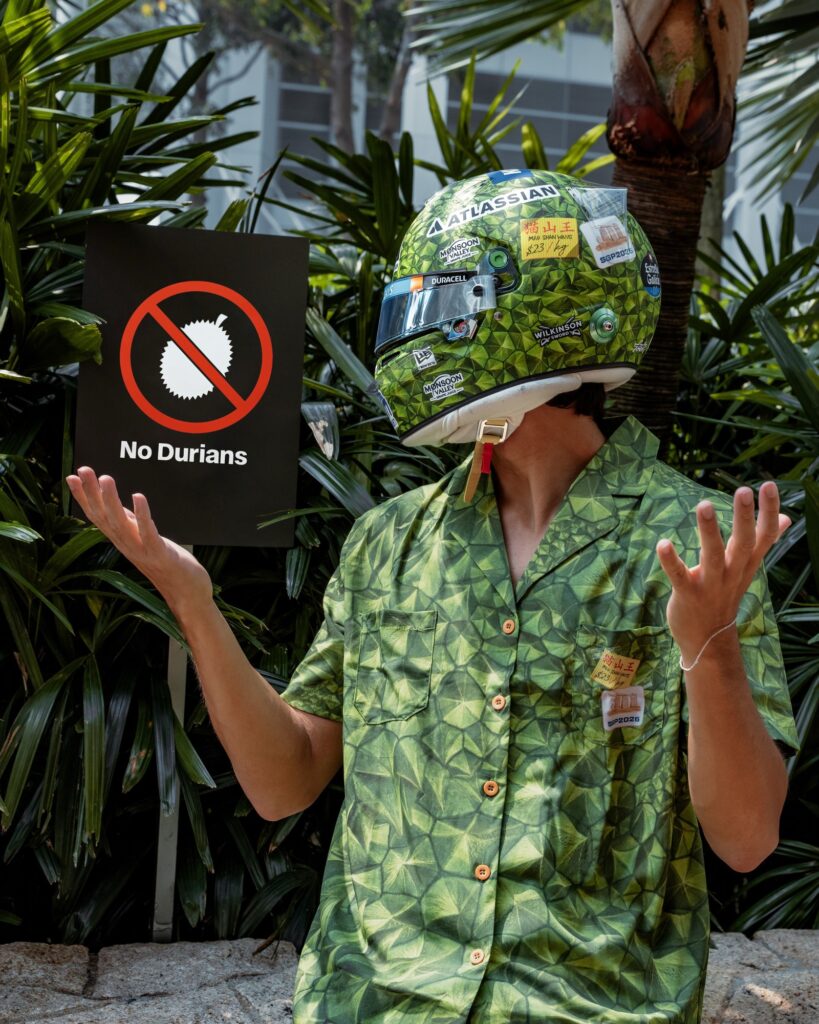 Alex Albon wearing his green durian-skin Singapore GP helmet and a matching shirt beside a No Durians sign