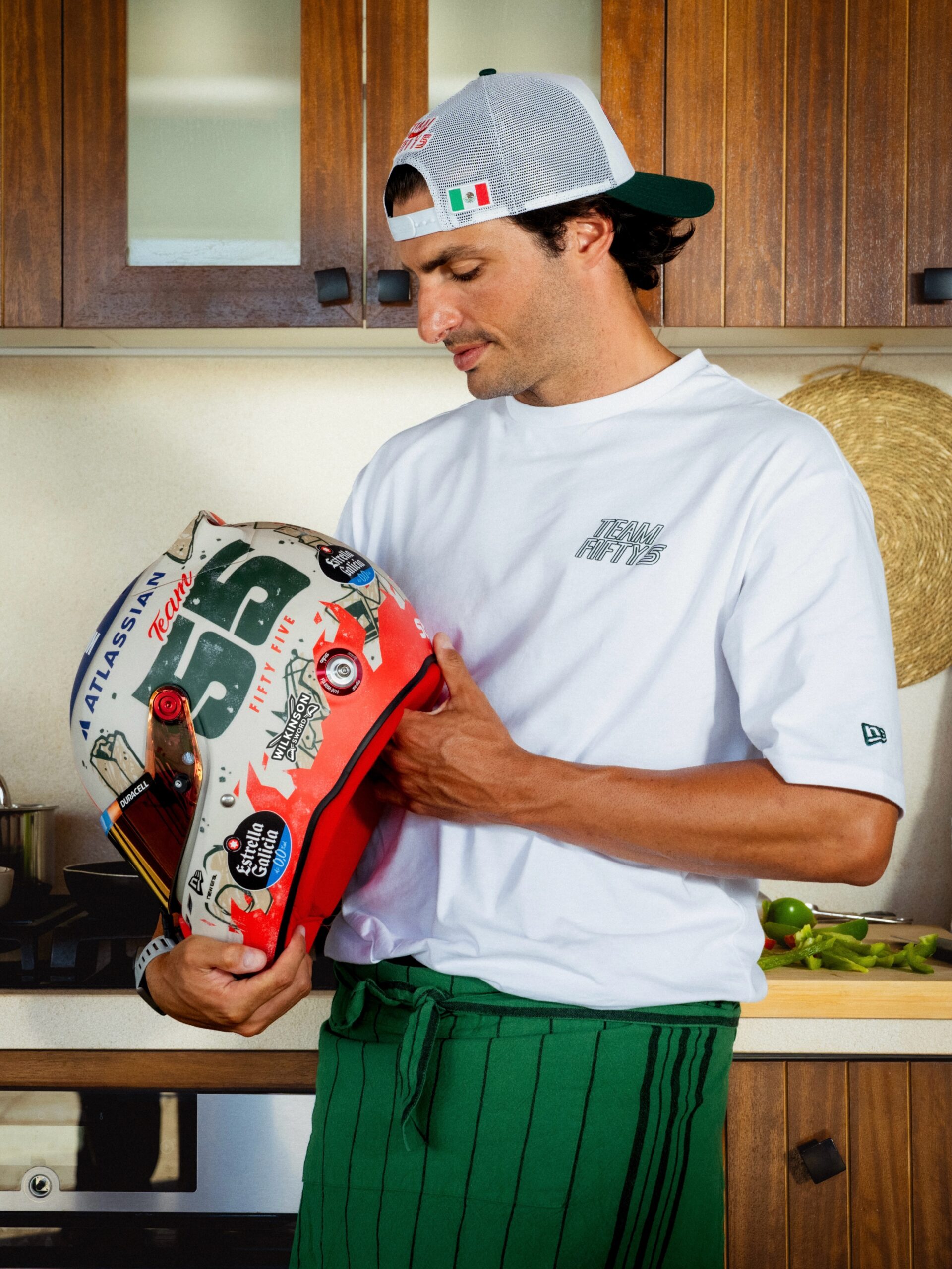 Carlos Sainz in a kitchen holding his 2026 Mexico City GP helmet, with ATLASSIAN, the green 55 and FIFTY FIVE