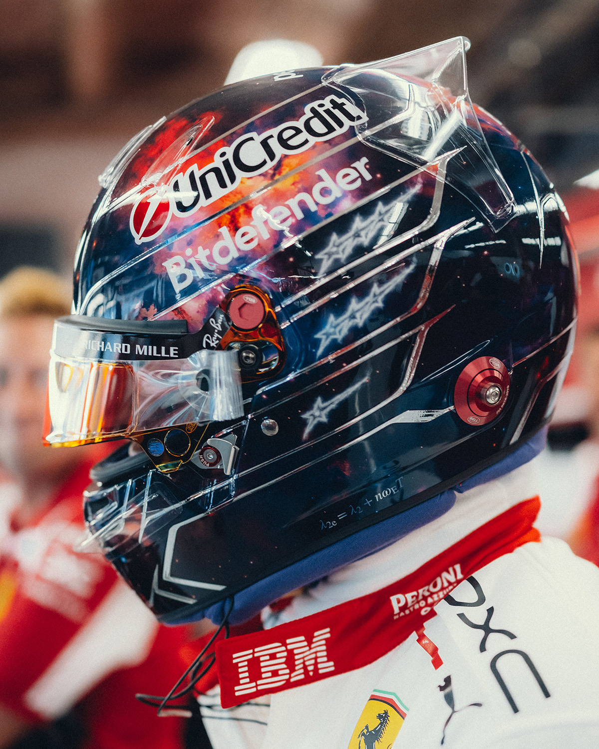 Lewis Hamilton's Austin 2026 helmet in profile: navy space scene with red and orange nebula, sketched white stars, chrome lines, gold mirrored visor