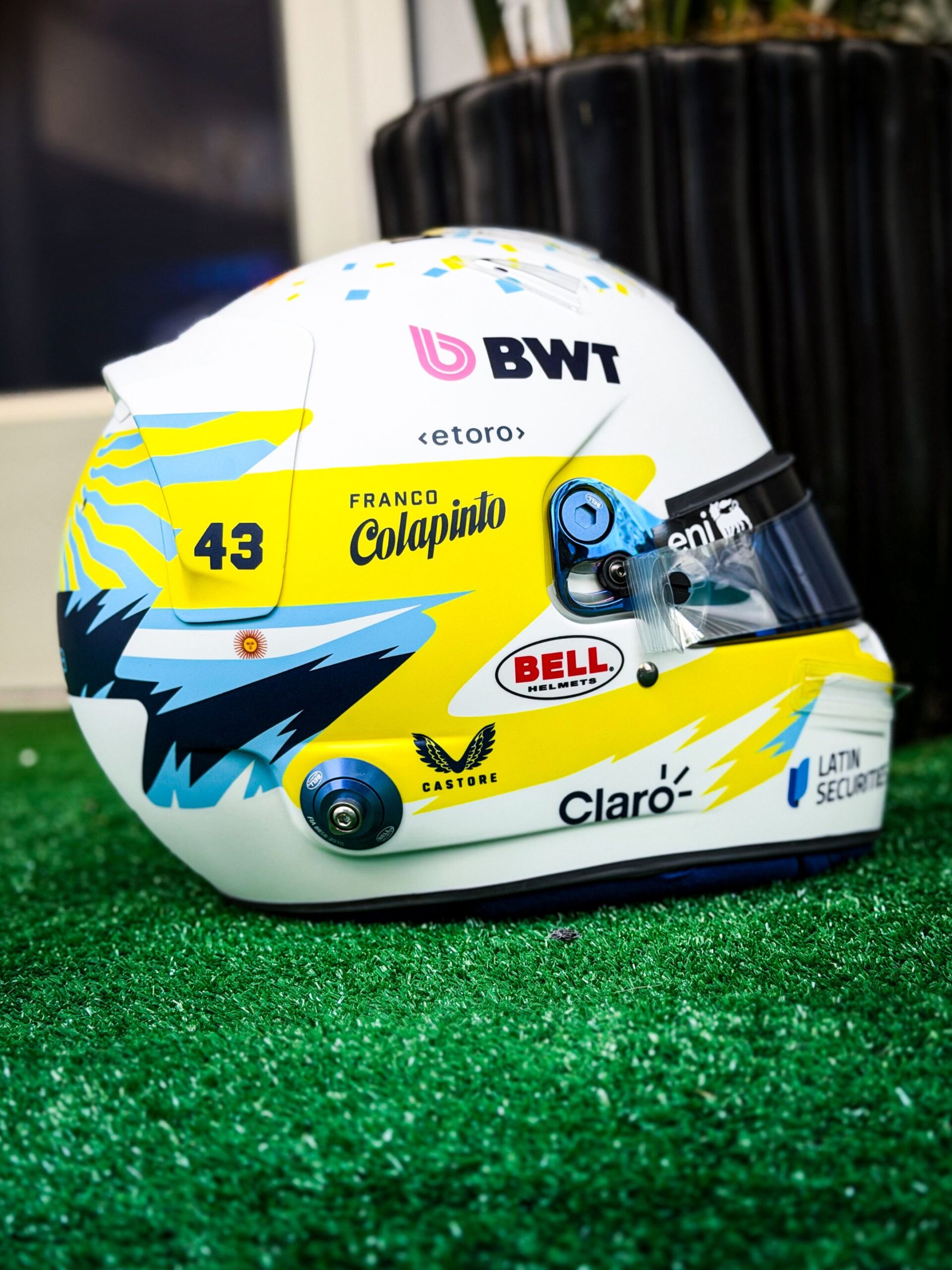Franco Colapinto's finished Austin 2026 helmet on artificial grass in the paddock: yellow band with FRANCO Colapinto and 43, light-blue and yellow rays
