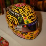 Three-quarter front view of Lando Norris's yellow 2026 Singapore GP helmet with black blobs, pink lotus flowers, Monster Energy and OKX