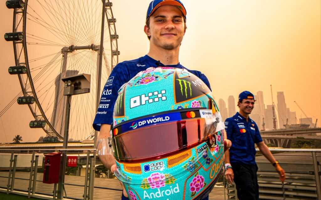 Oscar Piastri holding his turquoise 2026 Singapore GP helmet with beaded flowers in front of the Singapore Flyer